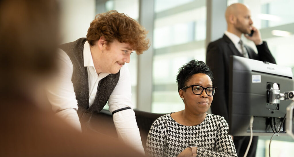 People working together at a computer workstation in an office, with another person using a phone in the background.