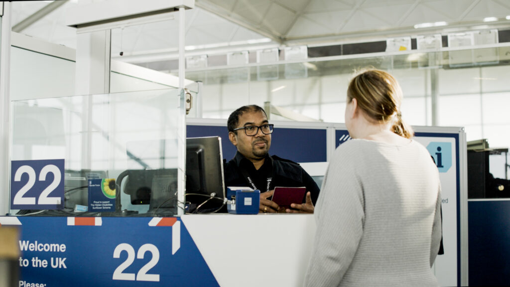 Border Control colleague standing behind a screen performing a passport check for a member of the public.