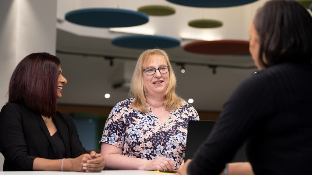 Three female colleagues sitting around a table, one has a laptop. 