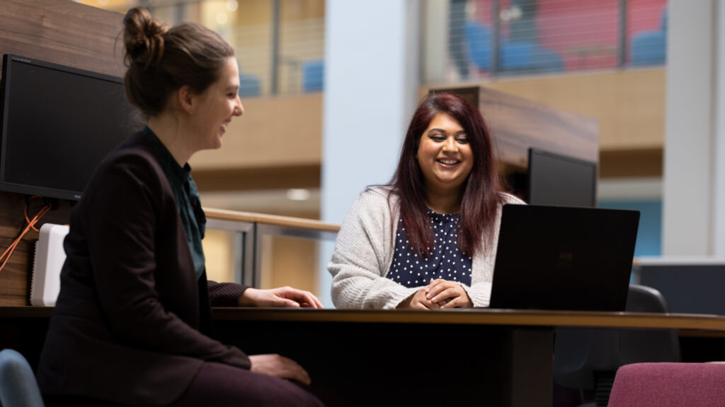 Two people seated at a table, looking toward a laptop.