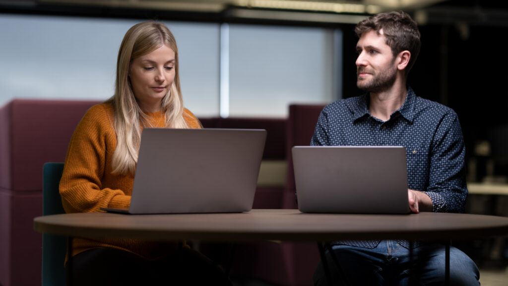 Two people seated at a round table in an indoor workspace, each using a laptop.
