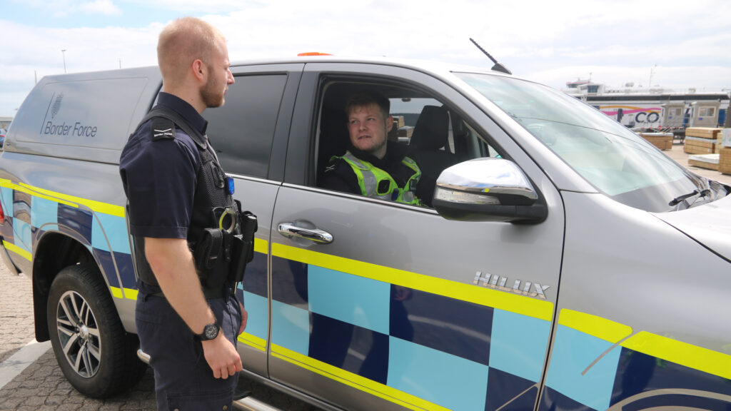 Outdoor setting showing one person in uniform standing beside a Border Force marked vehicle and another person in uniform seated inside.
